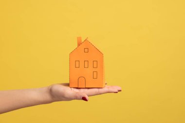 Real estate insurance, repairing services. Closeup of woman hand holding small orange paper house, concept of dream home purchase, mortgage. Indoor studio shot isolated on yellow background.