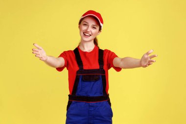 Mutlu, gülümseyen, elleri açık, kucaklaşmaya davet edilen, kollarıma gelen, tulumlu ve kırmızı şapkalı bir kadın. Sarı arka planda kapalı stüdyo çekimleri.