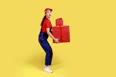 Side view portrait of hard working courier woman holding heavy presents boxes, looking at camera, door-to-door delivery, wearing overalls and red cap. Indoor studio shot isolated on yellow background.