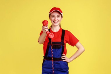Cep telefonunu tutan, onu arayıp sipariş veren, kameraya dost canlısı bakan, tulumlu ve kırmızı şapkalı bir kadının portresi. Sarı arka planda kapalı stüdyo çekimleri.