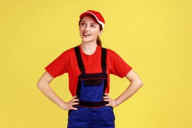 Pozitif hayalperest bir kadının portresi, elleri kalçasında, başka tarafa bakıyor, hoş bir şey düşünüyor, tulum giyiyor ve kırmızı şapka takıyor. Sarı arka planda kapalı stüdyo çekimleri.