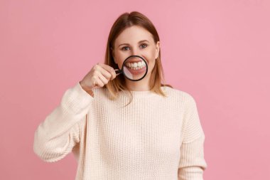 Portrait of positive attractive blond woman standing, holding magnifying glass on his teeth, looking at camera, wearing white sweater. Indoor studio shot isolated on pink background.