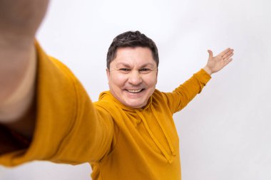 Middle aged handsome man taking selfie, looking at camera POV and showing welcome gesture, point of view of photo, wearing urban style hoodie. Indoor studio shot isolated on white background.