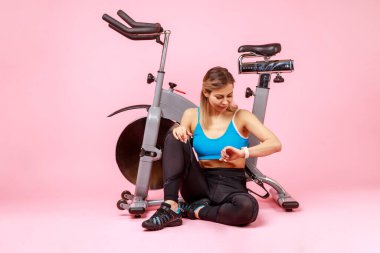 Full length portrait of athletic woman looking at her fitness tracker, checking indicators after workout, wearing sports tights and top. Indoor studio shot isolated on pink background.
