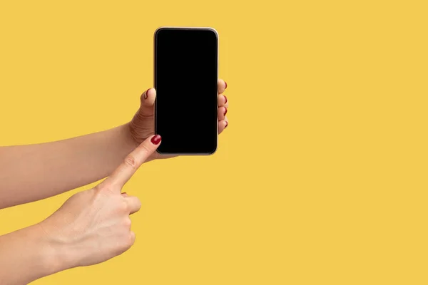 女性の手を保持し スマートフォンを表示し 空のディスプレイを指してのプロフィール側のビューを閉じます 黄色の背景に隔離された屋内スタジオショット — ストック写真