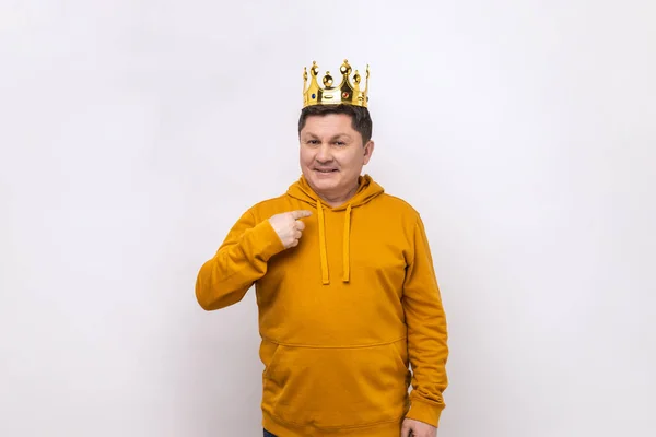 I\'m king. Middle aged man in golden crown and pointing himself, looking at camera with smile, superior privileged status, wearing urban style hoodie. Indoor studio shot isolated on white background.