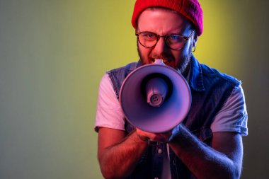 Angry hipster man holding megaphone near mouth loudly speaking, screaming, making announcement, paying attention at social problems. Indoor studio shot isolated on colorful neon light background.