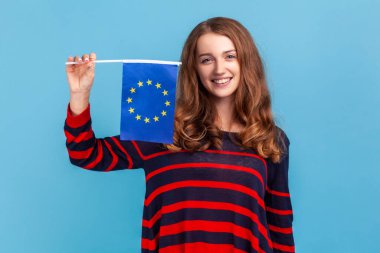 Optimistic woman wearing striped casual style sweater, showing flag of European union, looking at camera with smile, EU association and community. Indoor studio shot isolated on blue background.