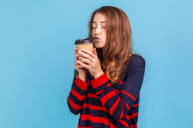 Attractive woman wearing striped casual style sweater, standing holding coffee to go, keeps lips pout, needs energy, happy to drink hot beverage. Indoor studio shot isolated on blue background.