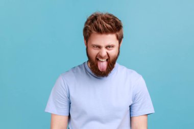 Ağzı açık kameraya bakan, çocuksu ve edepsiz yüz ifadesi takınan aptal, sakallı bir gencin portresi. Mavi arka planda kapalı stüdyo çekimi.