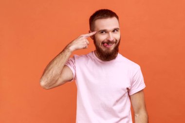 Portrait of bearded man showing stupid gesture, looking at camera with condemnation and tongue out, dumb suggestion, wearing pink T-shirt. Indoor studio shot isolated on orange background.