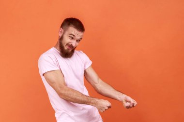 Güçlü sakallı bir adamın portresi yumrukları havada tutuyor, görünmez bir ip tutuyor ve objeyi çekiyor, gayretle çabalıyor, pembe tişört giyiyor. Turuncu arka planda kapalı stüdyo çekimleri.