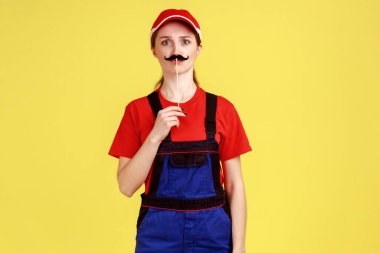 Elinde kağıt bıyıkla dikilip kameraya bakarken tulum ve kırmızı şapkayla eğlenen komik bir kadının portresi. Sarı arka planda kapalı stüdyo çekimleri.