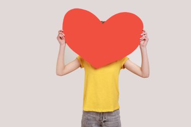 Anonymous date. Portrait of unknown girl in yellow casual style T- shirt hiding her face behind big read paper heart, holding symbol of love. Indoor studio shot isolated on gray background.