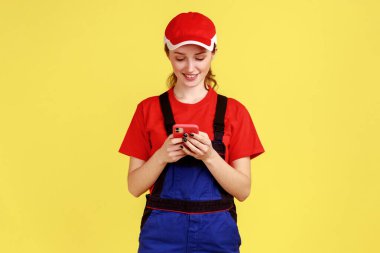 Çevrimiçi destek için cep telefonu kullanan genç ve gülümseyen kadın portresi, olumlu duygular ifade ediyor, tulum giyiyor ve kırmızı başlık takıyor. Sarı arka planda kapalı stüdyo çekimleri.