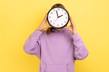 Portrait of young woman holding wall clock hiding her face, time management, schedule and meeting appointment, wearing purple hoodie. Indoor studio shot isolated on yellow background.