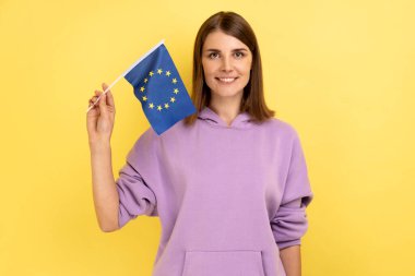 Patriotic woman with dark hair holding eu flag in hand looking at camera with toothy smile, patriotism, flag day celebration, wearing purple hoodie. Indoor studio shot isolated on yellow background.