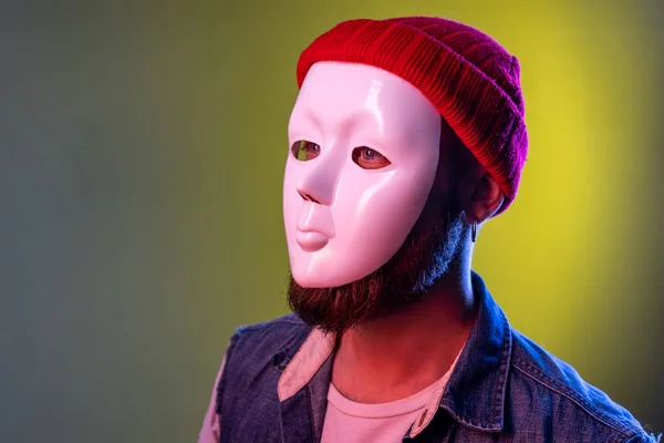 Side view of anonymous hipster man in white mask on face, hiding his identity, spying undercover, corruption, wearing hat and denim vest. Indoor studio shot isolated on colorful neon light background.