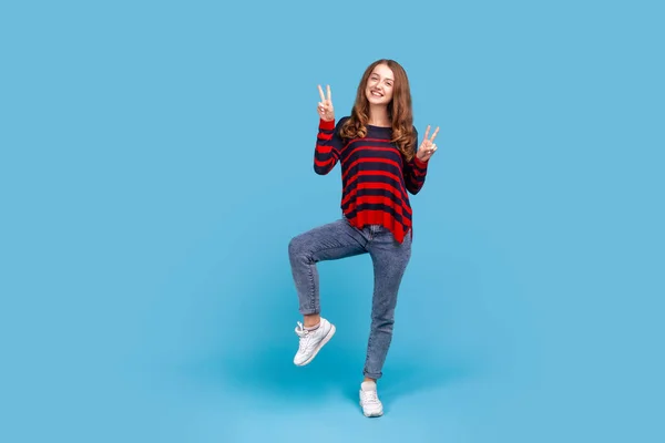 平和のサインを示す縞模様のカジュアルなスタイルのセーターを着て幸せな女性の完全な長さの肖像画 Vジェスチャーや笑顔 勝利を祝う 青い背景に隔離された屋内スタジオショット — ストック写真