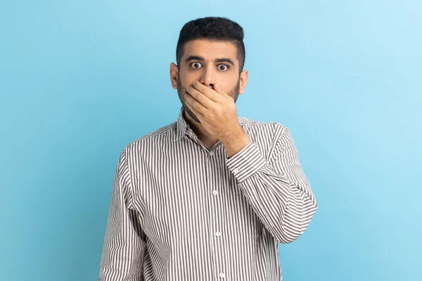 I won't tell. Portrait of amazed businessman covering mouth with hand, keeping secret, terrified with shocking news, wearing striped shirt. Indoor studio shot isolated on blue background.