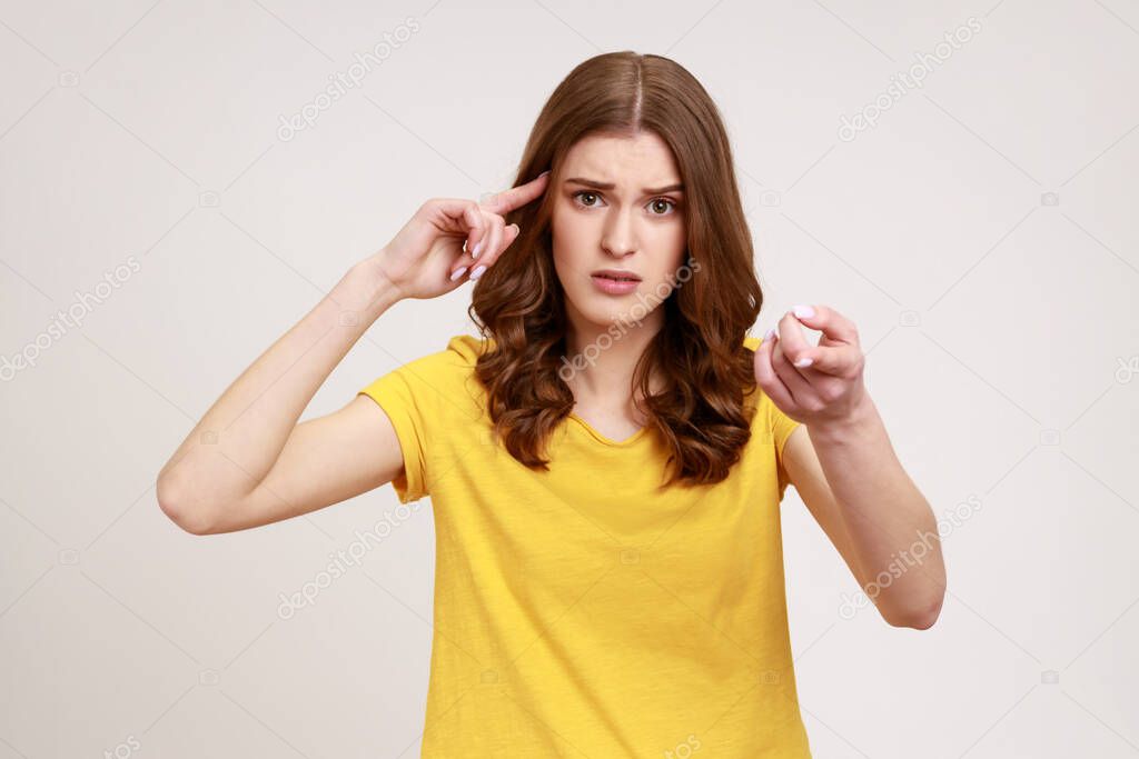 You are stupid! Self confident egoistic teenager girl wearing yellow T-shirt holding finger near temple and pointing on you, showing stupid gesture. Indoor studio shot isolated on gray background.