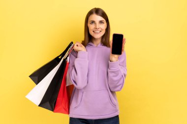 Tatmin olmuş bir şekilde ayakta duran mutlu kadın alışveriş torbaları ve reklam için boş ekranlı, mor kapüşonlu akıllı telefon gösteriyor. Sarı arka planda kapalı stüdyo çekimleri.
