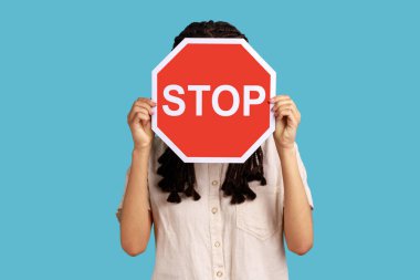 Unknown woman with black dreadlocks hiding her face behind red stop traffic symbol, warning, avoids forbidden actions, wearing white shirt. Indoor studio shot isolated on blue background.