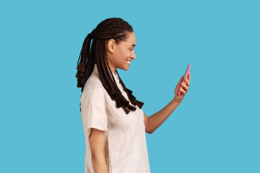 Kadının yan görüntüsü cep telefonuyla mesaj gönderiyor, internette tatil için hediyeleri arıyor, akıllı telefon uygulaması kullanıyor, web sayfasına göz atıyor. Mavi arka planda kapalı stüdyo çekimi.