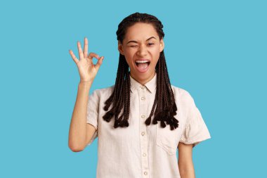 Siyah rastalı neşeli genç bir kadının portresi her şeyin yolunda olduğunu gösteriyor, mutlu görünüyor, beyaz gömlek giyiyor. Mavi arka planda kapalı stüdyo çekimi.