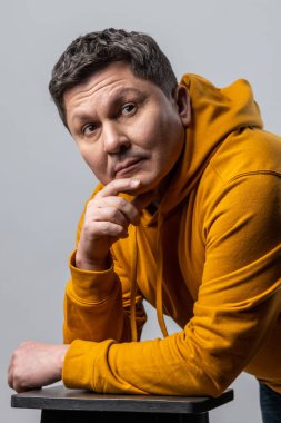 Düşünceli orta yaşlı bir adamın portresi, elleriyle sandalyeye yaslanmış, başka yere bakıyor, düşünceliliğini ifade ediyor, kentsel kapüşonlu kapüşonlu giyiyor. Beyaz arka planda izole edilmiş kapalı stüdyo görüntüleri.