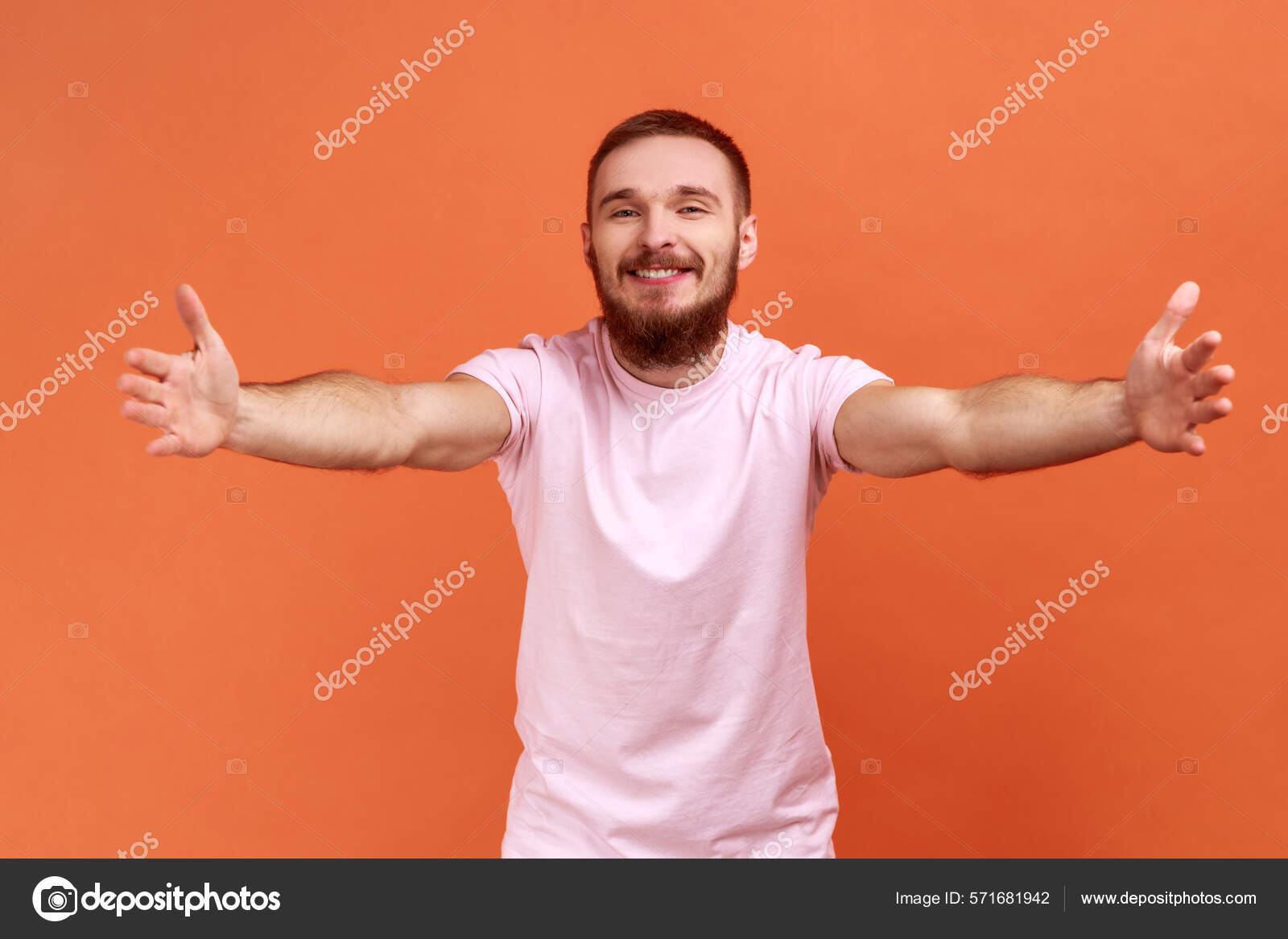 到我怀里来一个快乐的积极的留着胡子的男人向镜头伸出手来张开双臂拥抱你穿着粉色T恤的画像在橙色背景下被隔离的室内拍摄—