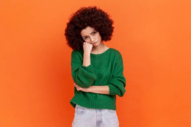 Afro saç stili, yeşil bir süveter giyen, çenesinin altında yumruğuyla sıkıcı bir hikaye dinleyen sıkılmış bir kadının portresi. Turuncu arka planda kapalı stüdyo çekimleri.