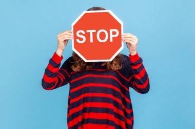 Bilinmeyen bir kadın, çizgili bir süveter giyiyor, yüzünü Dur sembolüyle kapatıyor, elinde kırmızı bir işaret, tehlike uyarısı, kısıtlama ve sınırlar var. Mavi arka planda kapalı stüdyo çekimi.