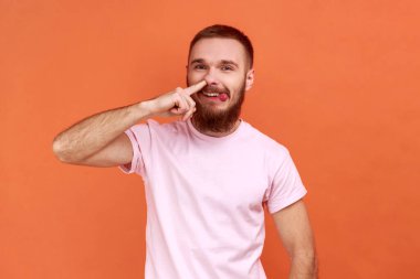 Portrait of bearded man standing, picking nose and sticking out tongue with dumb comical expression, fooling around, wearing pink T-shirt. Indoor studio shot isolated on orange background.