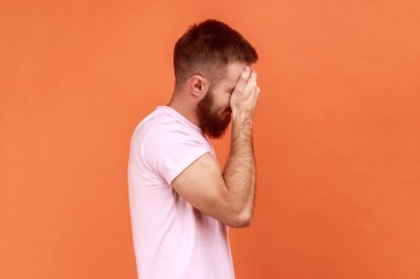 Genç, sakallı, sakallı bir adam bunu görmek istemiyor. Sorunları görmezden geliyor, stresli durumlardan saklanıyor, pembe tişört giyiyor. Turuncu arka planda kapalı stüdyo çekimleri.