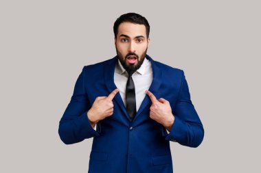 Şaşırmış sakallı adam kendini işaret ediyor, şaşırmış bir ifadeyle bakıyor, şok olduğunu söylüyor, inanılmaz bir başarı, resmi stil kıyafeti giyiyor. Gri arka planda izole edilmiş kapalı stüdyo görüntüleri.