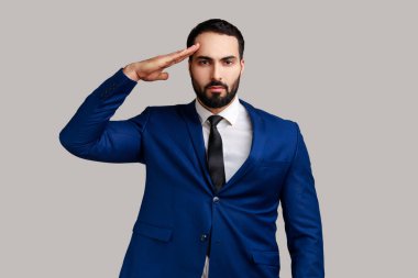 Ciddi derecede sıkı sakallı bir adamın portresi. Başının yanında el ele tutuşuyor, selam veriyor, onur ve zafer veriyor, askerileştiriyor, resmi bir tarz giyiyor. Gri arka planda izole edilmiş kapalı stüdyo görüntüleri.