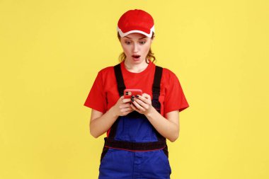 Online destek için cep telefonu kullanan, olumlu duygularını ifade eden, tulum giyen ve kırmızı şapka takan genç ve şok olmuş kadın portresi. Sarı arka planda kapalı stüdyo çekimleri.