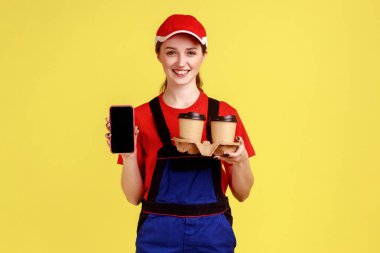 Elinde kahve taşıyan teslimatçı kadın, reklam için boş ekranlı cep telefonunu gösteriyor, kameraya bakıyor, tulum giyiyor ve kırmızı şapka takıyor. Sarı arka planda kapalı stüdyo çekimleri.
