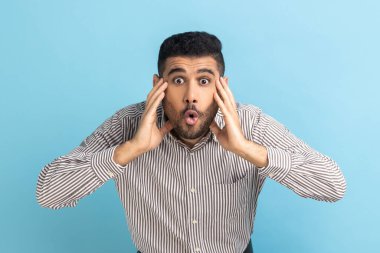 Vay canına, inanılmaz. Şaşırmış işadamı kameraya bakıyor ağzı açık ve kocaman gözleri var, kollarını kaldırıyor, çizgili gömlek giyiyor. Mavi arka planda kapalı stüdyo çekimi.
