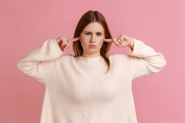 Bunu daha fazla duymak istemiyorum. Mutsuz sarışın kadının portresi ayakta duruyor ve parmağını kulaklarına sokuyor, kameraya bakıyor, beyaz kazak giyiyor. Pembe arka planda kapalı stüdyo çekimleri.