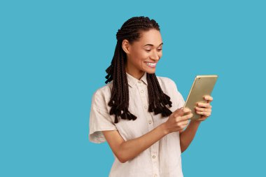 Siyah bukleli yaratıcı gülümseyen kadın ilerideki projelerde sosyal ağları kontrol etmek için tablet kullanıyor, beyaz gömlek giyiyor. Mavi arka planda kapalı stüdyo çekimi.