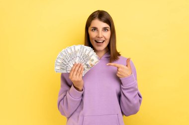 İyimser gülümseyen kadın elindeki dolar banknotlarını işaret ediyor, olumlu bir ifadeyle, büyük kârla, mor kapüşonlu bir şey giyiyor. Sarı arka planda kapalı stüdyo çekimleri.