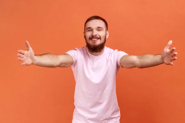 到我怀里来一个快乐的 积极的 留着胡子的男人向镜头伸出手来 张开双臂拥抱你 穿着粉色T恤的画像 在橙色背景下被隔离的室内拍摄 — 图库照片