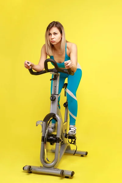 運動自転車で心臓トレーニングを行う美しいブロンドの女性の完全な長さ 体重を減らしたり 彼女の体の形を改善 青いスポーツウェアを身に着けています 黄色の背景に隔離された屋内スタジオショット — ストック写真