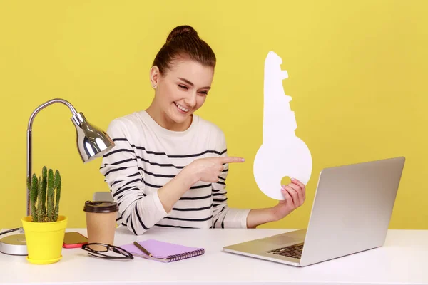 大きな紙キーを指し 職場に座っている間におもちゃの笑顔でノートパソコンの画面を見て女性のオフィスワーカー 家の購入の概念 黄色の背景に隔離された屋内スタジオショット — ストック写真
