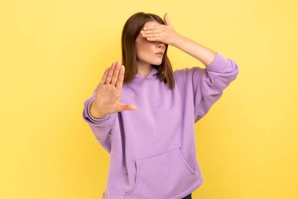 Don't want to look. Portrait of woman keeping hand on eyes and avoiding watch at something shameful, showing stop ban gesture, wearing purple hoodie. Indoor studio shot isolated on yellow background.