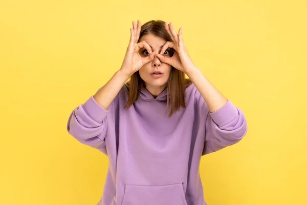 双眼鏡の手のジェスチャーを見て 驚きを表現し 紫色のパーカーを着て 指でメガネの形を作る驚きの女性 黄色の背景に隔離された屋内スタジオショット — ストック写真