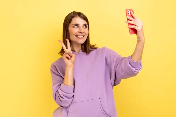 スマホに手を振って自撮りしながら ビデオ通話をしながらVサインを見せながら紫のパーカーを着て楽観的な幸せな女性の肖像画 黄色の背景に隔離された屋内スタジオショット — ストック写真