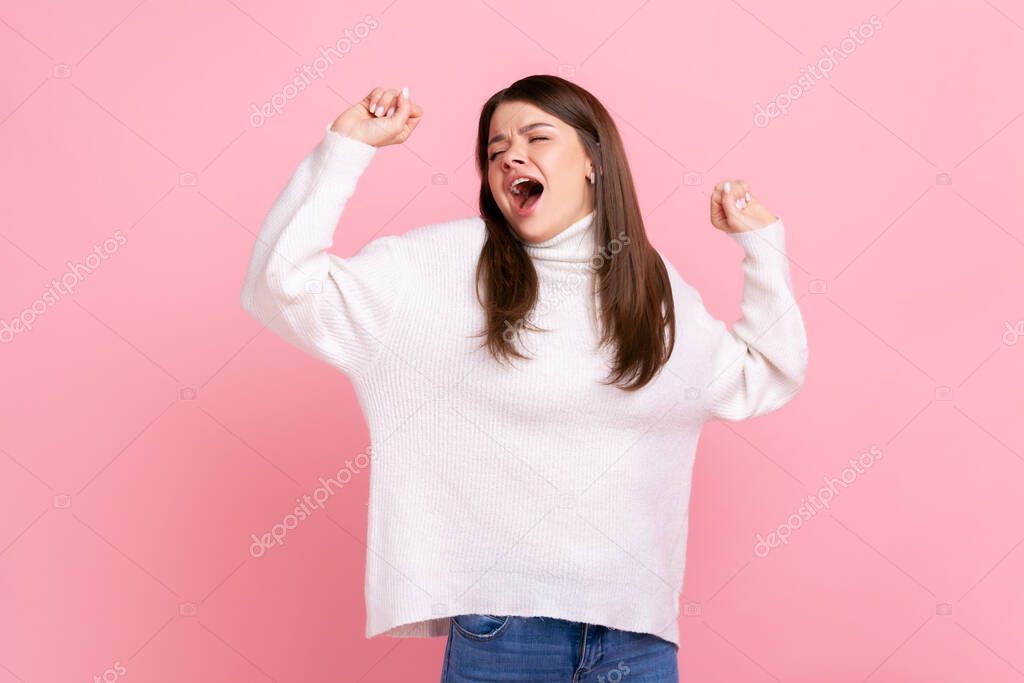 Sleepy young woman standing yawning with closed eyes, stretching arms, feeling exhausted need rest, wearing white casual style sweater. Indoor studio shot isolated on pink background.